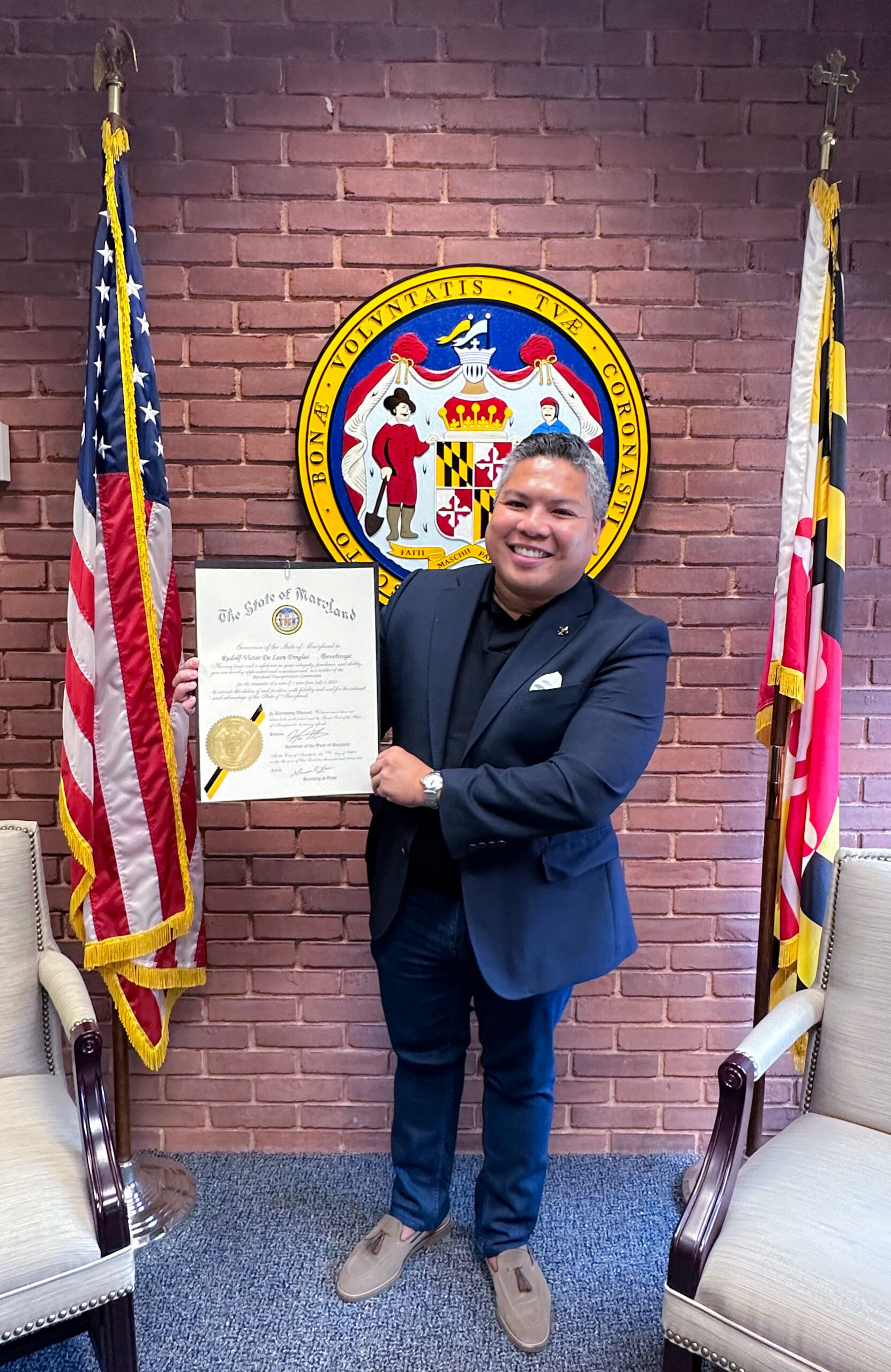 A person standing between the US flag and the Maryland flag holds up a certificate.